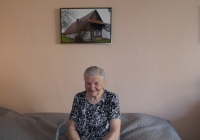 Zdenka Nováková v roce 2024, v pozadí fotografie rodného domu ve Vidlaté Seči