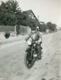 Josef Prejda na své motorce ČZ 125 jedoucí po hlavní silnici v Uhlířově v úrovni dvorku Frankových. Dům v pozadí je původně Prejdových, později Wolfových. Fotografie z 50. let 20. století
